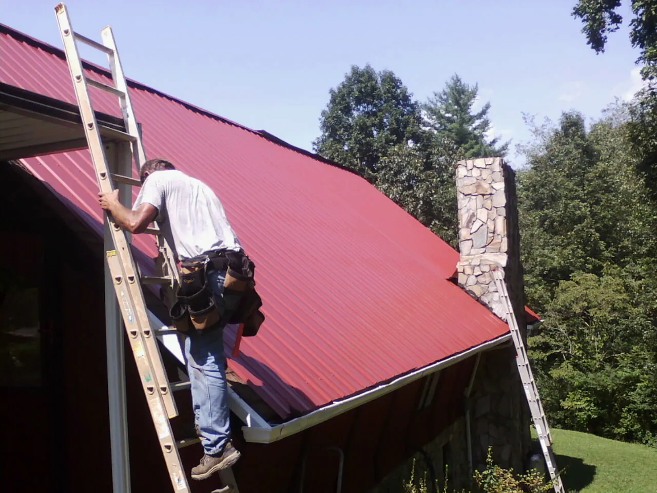 Roofer on a ladder beside a red metal roof and stone chimney — Soffit & Fascia Repair by Roofing Brothers in Colerain